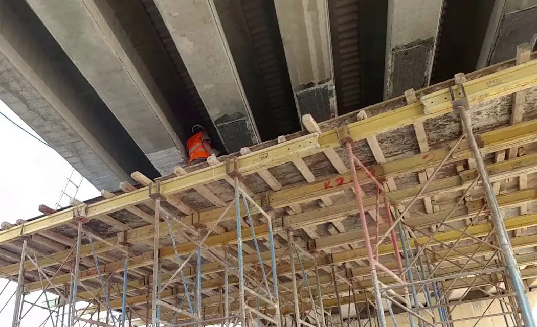 Otoyoldaki bir köprünün öngermeli kirişleri alttan görülüyor; kirişlerin altına iskele kurulmuş, kiriş uçları karbon fiberle sarılmış, kirişlerin arasında turuncu yelekli bir usta çalışıyor.